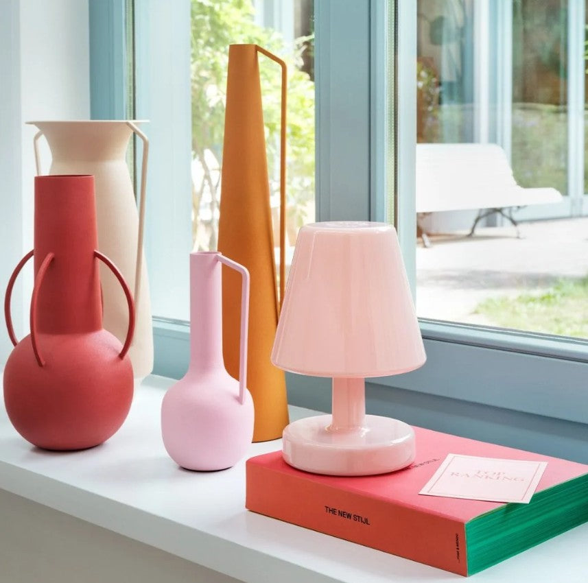 A pink Edison the Ambiance table lamp with a conical shade and round base sits atop a red book on a windowsill, surrounded by sculptural vases in red, cream, pink, and orange.